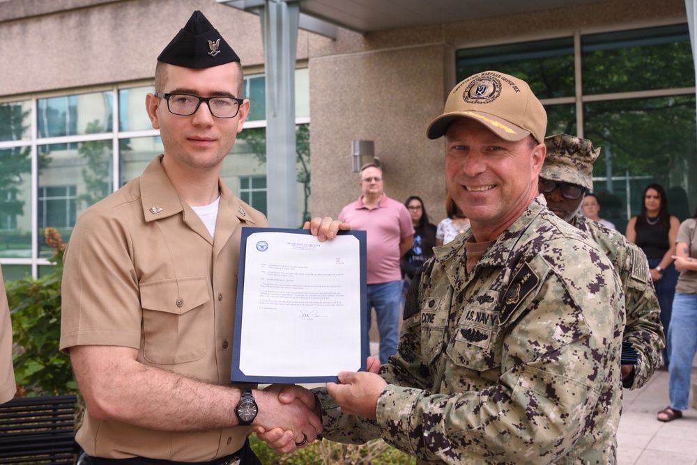 Two U.S. Navy personnel are shaking hands while jointly holding an official document with a Department of Defense seal, suggesting a formal recognition or award ceremony. The younger sailor on the left is in service khaki uniform with a garrison cap, while the senior officer on the right wears NWU Type III camouflage and a tan command ball cap. Several civilians and another uniformed service member are visible in the background observing the event.