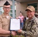 Two U.S. Navy personnel are shaking hands while jointly holding an official document with a Department of Defense seal, suggesting a formal recognition or award ceremony. The younger sailor on the left is in service khaki uniform with a garrison cap, while the senior officer on the right wears NWU Type III camouflage and a tan command ball cap. Several civilians and another uniformed service member are visible in the background observing the event.