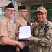 A U.S. Navy officer in digital camouflage (NWU Type III) is presenting a certificate or commendation document to a junior sailor in khaki uniform during what appears to be an outdoor military ceremony. The officer's name tape reads 'CONE' and he is wearing a U.S. Navy cap along with what appears to be a senior enlisted or officer rank insignia. A second sailor in khaki uniform stands in the background observing the presentation.