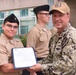 A U.S. Navy officer in digital camouflage (NWU Type III) uniform and a tan cap presents a certificate or official document to a junior sailor in service khaki uniform. The ceremony appears to be taking place outdoors in front of a modern building, with additional sailors in formation visible in the background. This appears to be an awards or recognition ceremony.