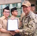 A U.S. Navy officer in digital camouflage NWU (Navy Working Uniform) is presenting a certificate or official document to a junior sailor in tan service uniform during what appears to be a formal recognition ceremony. The event is taking place outdoors in front of a building, with additional uniformed personnel and civilians visible in the background. The officer's cap bears insignia consistent with a Naval Warfare Group unit.