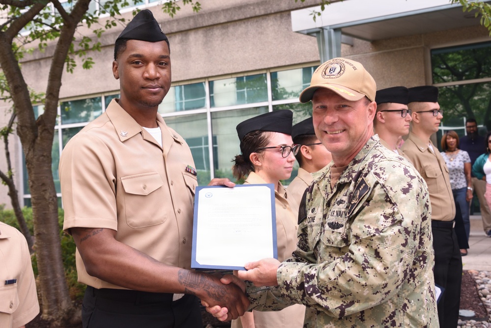 A U.S. Navy ceremony is taking place outdoors in front of a modern building. A senior Navy officer in digital camouflage uniform and tan cap is presenting a certificate or award document to a junior sailor in tan service uniform. Several other sailors in uniform are standing in formation in the background.