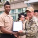 A U.S. Navy ceremony is taking place outdoors in front of a modern building. A senior Navy officer in digital camouflage uniform and tan cap is presenting a certificate or award document to a junior sailor in tan service uniform. Several other sailors in uniform are standing in formation in the background.