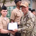 A U.S. Navy officer in digital camouflage (NWU Type III) uniform and tan cap is presenting a certificate or award to a junior sailor in khaki service uniform during what appears to be an outdoor military ceremony. Several other sailors in similar khaki uniforms and black garrison caps are visible in the background. The setting appears to be an outdoor courtyard or plaza near a building.