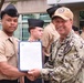 A U.S. Navy officer is presenting a certificate or official document to a junior enlisted sailor during what appears to be a formal military ceremony held outdoors. The officer, whose name tag reads 'CONE' and is wearing Navy Working Uniform (NWU) camouflage with a tan cap, is smiling broadly while shaking hands with the recipient. Several other sailors in tan service uniforms and garrison caps are visible in the background.