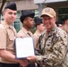 A U.S. Navy officer in digital camouflage (NWU Type III) uniform is presenting a certificate or commendation document to a junior sailor in tan service uniform during what appears to be an outdoor military ceremony. The presenting officer wears a tan cap with an insignia patch and has a name tape reading 'CONE' visible on his uniform. Multiple other sailors in tan uniforms and black garrison caps are visible in the background.