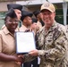 A U.S. Navy ceremony is taking place outdoors where a service member named 'CONE' is presenting an official document or certificate to a female service member in tan/khaki uniform. Both individuals are smiling and holding the document together for what appears to be a formal recognition or award presentation. Additional Navy personnel in uniform are visible in the background.