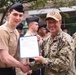 A U.S. Navy ceremony is taking place outdoors, where a senior officer identified by a name tape reading 'CONE' and wearing U.S. Navy digital camouflage (NWU) is presenting a framed certificate or commendation letter to a junior sailor in Service Khaki uniform. Both individuals are shaking hands while holding the document together. Several other Navy personnel in various uniforms are visible in the background.