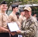 A U.S. Navy officer in digital camouflage (NWU Type I) is presenting a certificate or award document to a junior enlisted sailor in service khaki uniform during what appears to be a military ceremony. The officer's name tape reads 'CONE' and he is wearing a tan ball cap with a military insignia. Several other sailors in service khaki uniforms are standing in formation in the background.