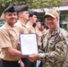 A U.S. Navy officer wearing NWU (Navy Working Uniform) with a tan cap is presenting a certificate or official document to a junior sailor in khaki uniform during what appears to be a formal recognition ceremony. The event takes place outdoors in front of a modern building, with a formation of sailors in khaki uniforms and black berets standing at attention in the background. The officer's name tape reads 'CONE' and his uniform displays 'U.S. NAVY' markings.