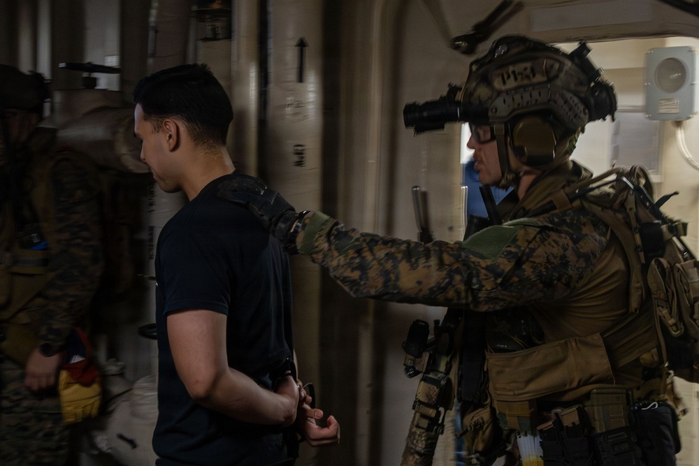 A fully equipped military operator in camouflage uniform and tactical gear, including a helmet with night-vision device, is escorting a restrained individual whose hands appear to be zip-tied or handcuffed behind their back. The setting appears to be the interior of a ship or large vessel, consistent with a maritime interdiction or boarding operation. Additional personnel in similar military attire are visible in the background.