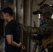 A fully equipped military operator in camouflage uniform and tactical gear, including a helmet with night-vision device, is escorting a restrained individual whose hands appear to be zip-tied or handcuffed behind their back. The setting appears to be the interior of a ship or large vessel, consistent with a maritime interdiction or boarding operation. Additional personnel in similar military attire are visible in the background.