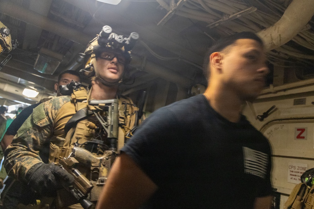 The image shows a heavily equipped military or law enforcement operator in full tactical gear, including night vision goggles mounted on a helmet, body armor, and a sidearm, moving through what appears to be the interior corridor of a naval vessel. A second individual in a black t-shirt with an American flag patch is visible in the foreground, slightly motion-blurred. The setting appears to be a maritime boarding or vessel search operation.