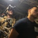 The image shows a heavily equipped military or law enforcement operator in full tactical gear, including night vision goggles mounted on a helmet, body armor, and a sidearm, moving through what appears to be the interior corridor of a naval vessel. A second individual in a black t-shirt with an American flag patch is visible in the foreground, slightly motion-blurred. The setting appears to be a maritime boarding or vessel search operation.