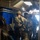 A group of heavily armed and equipped military personnel in full combat gear are staged inside what appears to be a naval vessel or military transport, moving toward a bright exit point. The soldiers are wearing multicam camouflage uniforms, tactical helmets, body armor, and carrying assault rifles. The setting suggests a maritime boarding operation or tactical insertion exercise.