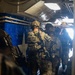 A group of heavily armed and equipped military personnel in full combat gear are staged inside what appears to be a naval vessel or military transport, moving toward a bright exit point. The soldiers are wearing multicam camouflage uniforms, tactical helmets, body armor, and carrying assault rifles. The setting suggests a maritime boarding operation or tactical insertion exercise.