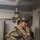 A fully equipped military service member, identifiable by MARPAT (Marine pattern) digital camouflage uniform, stands in what appears to be a naval vessel interior. The individual is wearing extensive tactical gear including a ballistic helmet with hearing protection, body armor, and a personal flotation device. They appear to be communicating via a radio or push-to-talk device held to their face.