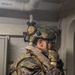 A fully equipped military service member, identifiable by MARPAT (Marine pattern) digital camouflage uniform, stands in what appears to be a naval vessel interior. The individual is wearing extensive tactical gear including a ballistic helmet with hearing protection, body armor, and a personal flotation device. They appear to be communicating via a radio or push-to-talk device held to their face.