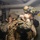 Two military personnel in full combat gear are positioned inside what appears to be a naval vessel corridor or compartment. Both are wearing camouflage uniforms, tactical helmets with night vision device mounts, and carrying tactical backpacks. The foreground soldier is adjusting equipment near his helmet while the background soldier appears to be aiming or observing through a scope or optic.