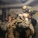 Two military personnel in full combat gear are positioned inside what appears to be a naval vessel corridor or compartment. Both are wearing camouflage uniforms, tactical helmets with night vision device mounts, and carrying tactical backpacks. The foreground soldier is adjusting equipment near his helmet while the background soldier appears to be aiming or observing through a scope or optic.
