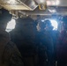 The image shows a group of military personnel inside what appears to be a naval vessel or military transport craft. Several individuals wearing tactical gear, helmets, and communication headsets are gathered near an open hatch or doorway with bright light streaming through. One person on the right is wearing a red vest, likely indicating a safety or crew role.