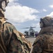 Two U.S. Marines in full combat gear observe a military helicopter on what appears to be a naval vessel's flight deck. The helicopter, likely a UH-1Y Venom or similar variant, is on the deck with rotor blades spinning while personnel gather around it. The scene suggests a pre-boarding or loading operation taking place at sea.