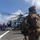 A military operation is underway on the flight deck of a naval vessel, with armed Marines in full combat gear approaching a helicopter marked '61' with its rotors spinning. Deck crew members in purple jerseys are visible near the aircraft, suggesting active flight deck operations. The scene appears to be a helicopter insertion or extraction exercise at sea.