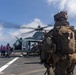 A military operation is underway on the flight deck of a naval vessel, with armed Marines in full combat gear approaching a helicopter marked '61' with its rotors spinning. Deck crew members in purple jerseys are visible near the aircraft, suggesting active flight deck operations. The scene appears to be a helicopter insertion or extraction exercise at sea.