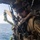 A military service member, identifiable by MARPAT (Marine pattern) camouflage uniform, is positioned at the open door of a helicopter over open ocean. The individual is equipped with full tactical gear including a flight helmet with NVG mount, communications headset, body armor, and multiple antennas. The image appears to capture a maritime helicopter operation, possibly a fast-rope or insertion exercise.