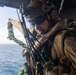 A military service member, identifiable by MARPAT (Marine pattern) camouflage uniform, is positioned at the open door of a helicopter over open ocean. The individual is equipped with full tactical gear including a flight helmet with NVG mount, communications headset, body armor, and multiple antennas. The image appears to capture a maritime helicopter operation, possibly a fast-rope or insertion exercise.