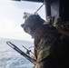 A military service member in full combat gear is positioned at the open door of a helicopter, looking out over a large body of open water. The individual is equipped with a tactical helmet, communications headset, camouflage uniform, body armor, and a backpack. The image appears to capture an airborne maritime operation or reconnaissance mission.