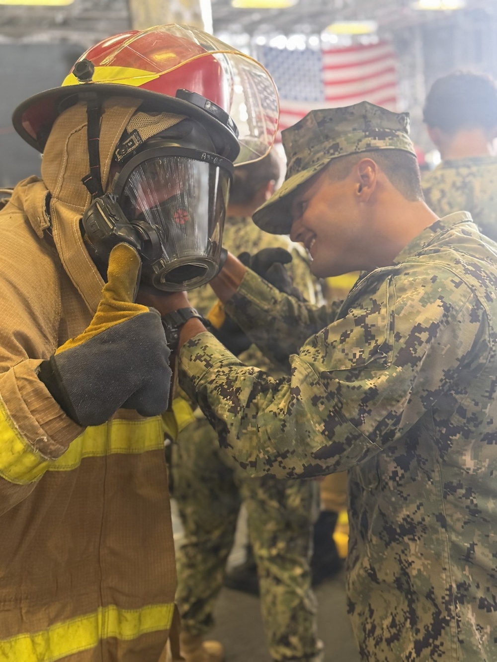 A U.S. Navy sailor in NWU Type III digital camouflage uniform assists another person in donning firefighting gear, including a full-face SCBA mask and structural firefighting helmet. The scene appears to be a damage control or firefighting training exercise, likely aboard a naval vessel or training facility. An American flag is visible in the background.