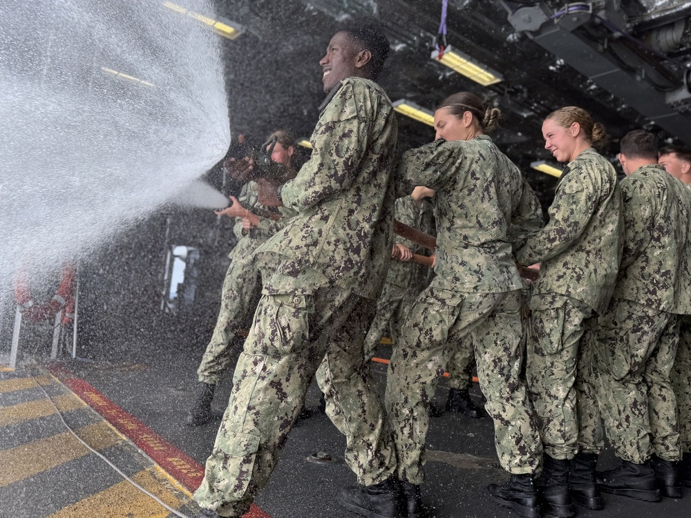 A group of U.S. Navy personnel in NWU Type III (digital camouflage) uniforms are conducting firefighting hose training, likely aboard a naval vessel. The lead sailor is directing a high-pressure water stream while teammates form a line behind, bracing against the hose's recoil. The atmosphere appears energetic and training-focused, with several personnel smiling.
