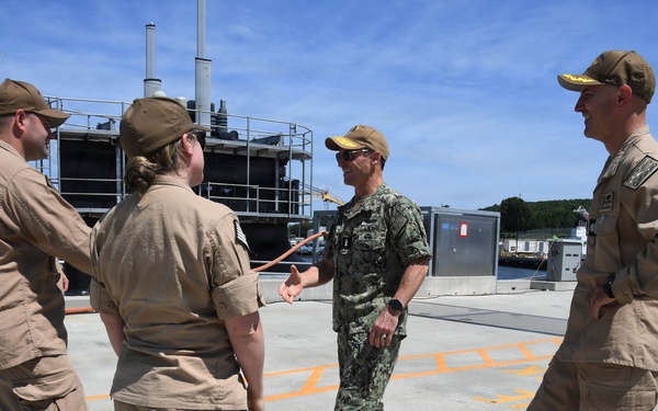 A group of five military personnel are gathered outdoors on what appears to be a naval installation or pier area. One individual wearing Navy Working Uniform (NWU) camouflage pattern is engaged in conversation with others dressed in tan/khaki utility uniforms. The setting includes industrial equipment and structures consistent with a naval facility.