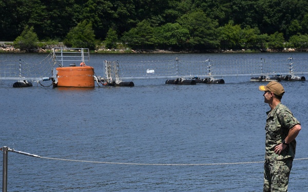 A military personnel member in camouflage uniform stands on top of a submarine hull at a naval facility, observing the surrounding waterway. The submarine is moored with various hoses and lines visible on its deck. In the background, floating barriers or boom systems are deployed across the water, with dense tree-covered hills beyond.