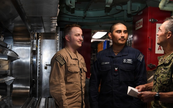 Three U.S. Navy personnel are engaged in conversation inside what appears to be a ship's galley or food service area. Two junior sailors in different working uniforms are speaking with a senior officer in camouflage NWU (Navy Working Uniform). The setting features stainless steel food service equipment and a red bulkhead with safety signage visible in the background.