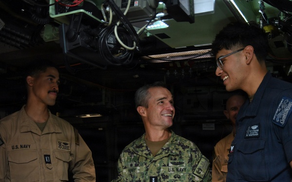 Three U.S. Navy personnel are engaged in a conversation inside what appears to be a submarine or naval vessel control room. The central figure, wearing a camouflage NWU (Navy Working Uniform) with what appears to be senior officer insignia, is smiling and interacting with a junior sailor in a dark navy uniform. The setting features complex overhead equipment, wiring, and instrumentation typical of a submarine interior.