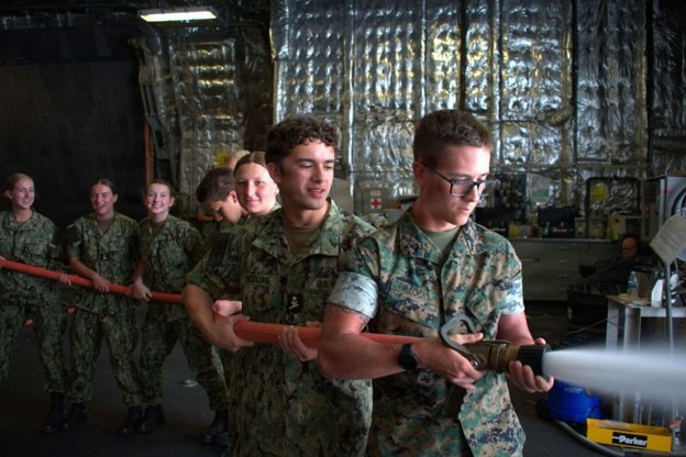 A group of military personnel in camouflage uniforms are conducting firefighting or damage control training inside what appears to be a ship or industrial facility. Two individuals in the foreground are operating an orange fire hose with a nozzle, while several others form a line behind them holding the hose. The background features reflective metallic insulation material typical of naval vessel interiors.