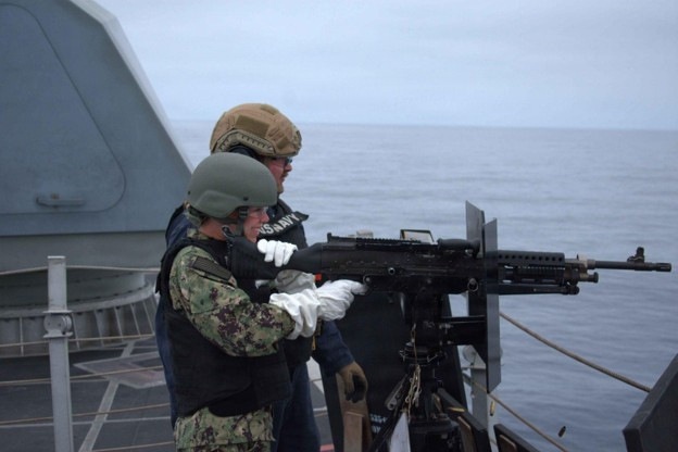 Two military personnel are positioned on the deck of a naval vessel, with one individual operating a mounted machine gun aimed toward the open sea. Both are wearing helmets and protective gear, suggesting a live-fire exercise or weapons qualification drill. The overcast sky and calm ocean provide the backdrop for what appears to be a routine naval training evolution.