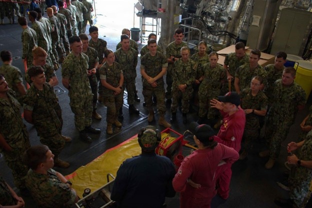 A group of military personnel in camouflage uniforms are gathered in what appears to be a ship's hangar bay or large interior deck space. Two instructors wearing red shirts are conducting a training demonstration, with a yellow stretcher/litter and medical equipment laid out on the deck floor. The personnel are arranged in a semicircle observing what appears to be a casualty care or medical evacuation training exercise.