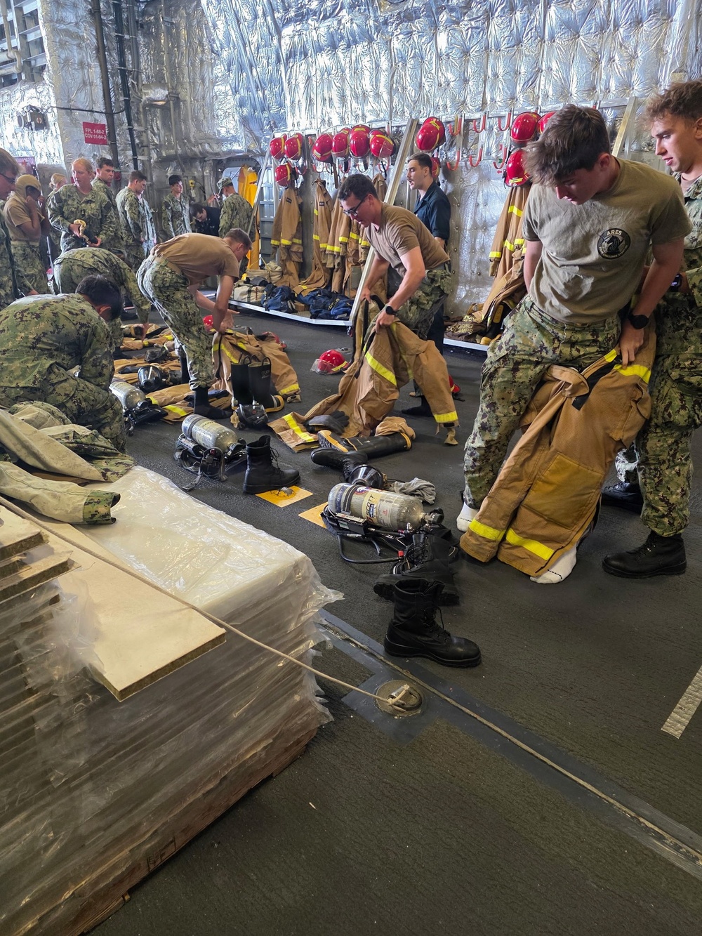 Military personnel, appearing to be U.S. Navy sailors in digital camouflage uniforms, are donning or handling firefighting gear in what appears to be a ship's interior compartment. The space is equipped with firefighting equipment including turnout gear, red helmets, SCBA (Self-Contained Breathing Apparatus) tanks, and boots laid out on the deck. This appears to be a damage control or firefighting drill conducted aboard a naval vessel.