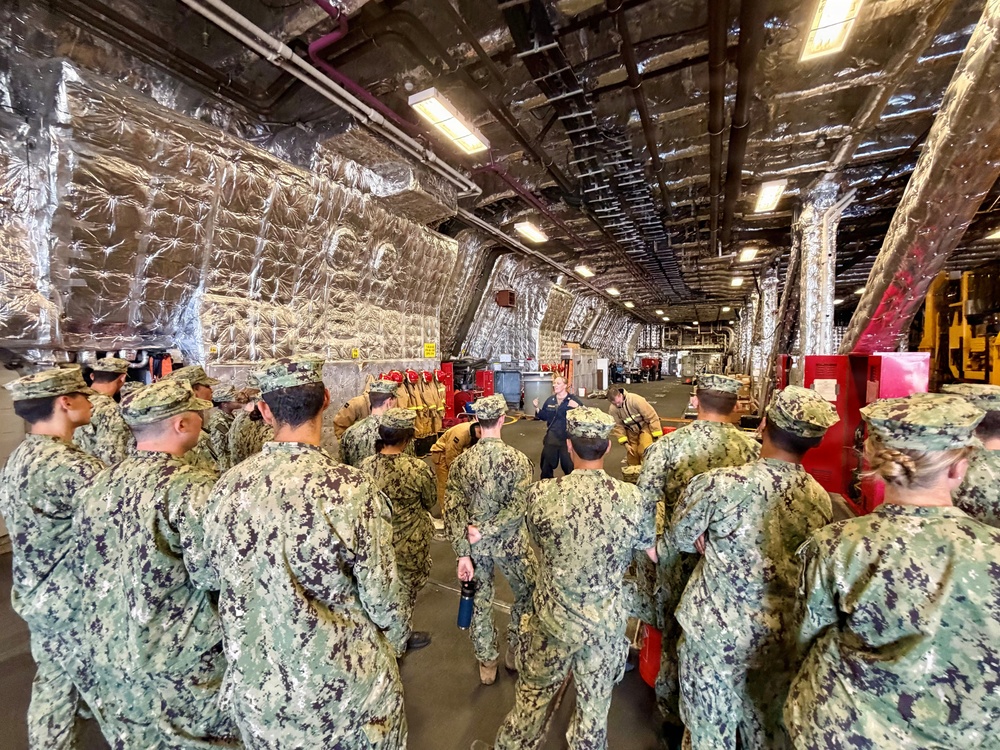 A large group of U.S. Navy personnel in NWU Type I (Navy Working Uniform) digital camouflage are gathered in what appears to be a ship's hangar bay or damage control training facility. An instructor in dark uniform stands at the front addressing the group, with firefighting equipment and gear visible in the background. The space features distinctive silver thermal/fire-resistant insulation lining the walls and ceiling, consistent with a naval vessel's interior.