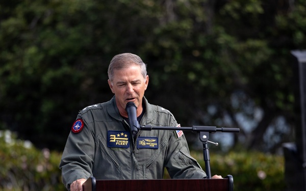 A military officer in a green flight suit stands at a wooden podium speaking into a microphone at an outdoor ceremony. The individual wears a 'Third Fleet' patch with three stars visible on their uniform, indicating a Vice Admiral rank. The setting appears to be a formal military outdoor event with landscaped grounds in the background.