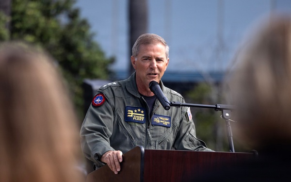 A military officer in a green flight suit is speaking at a podium with a microphone during what appears to be an outdoor ceremony or official event. The officer wears patches including one reading 'Third Fleet' and displays two stars on the collar, indicating the rank of Vice Admiral. Two audience members are visible in the foreground, blurred by shallow depth of field.