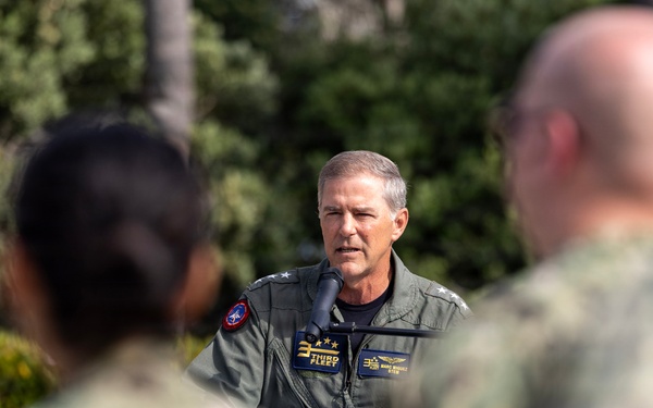 A senior U.S. Navy officer in a green flight suit is addressing a group of sailors in Navy Working Uniform (NWU) camouflage outdoors. The officer's flight suit displays a 'Third Fleet' patch and two-star rank insignia, indicating the rank of Vice Admiral. The setting appears to be a military installation with landscaped greenery in the background.