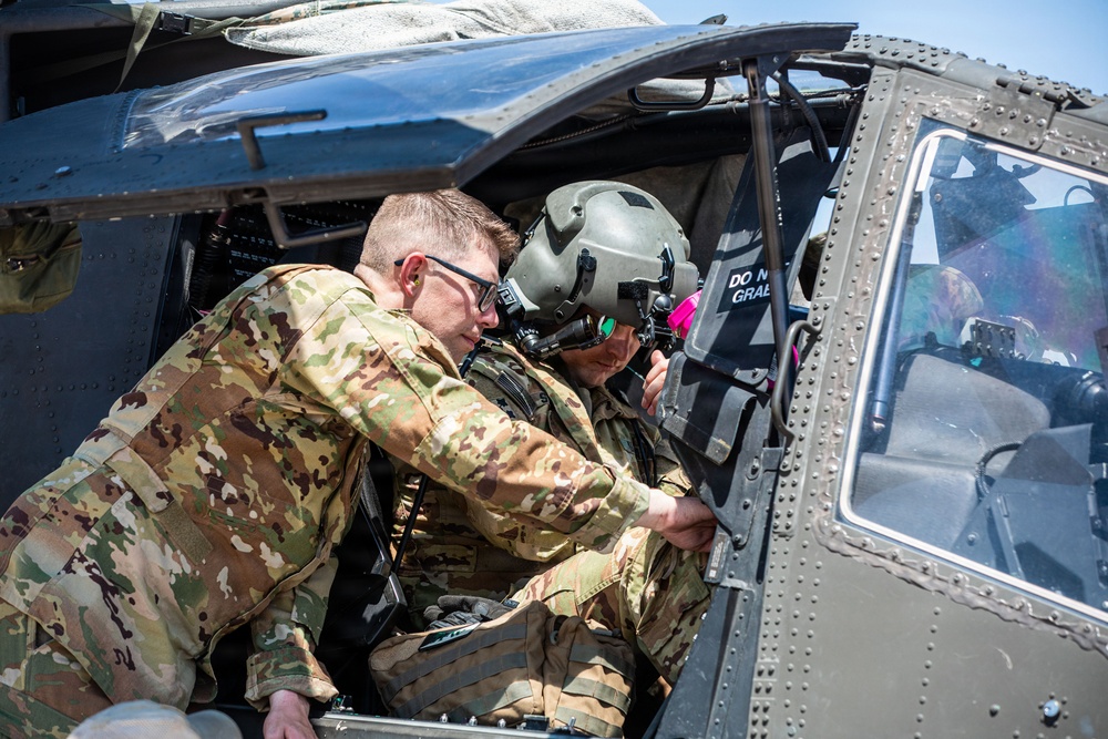 Two military personnel in camouflage uniforms are interacting near the open cockpit door of a military helicopter, likely a UH-60 Black Hawk. One individual wears a flight helmet with a visor, suggesting they are a pilot or aircrew member, while the other wears glasses and appears to be pointing at or adjusting something inside the cockpit. A third person is partially visible through the cockpit windshield.