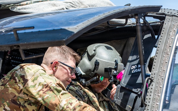 Two military personnel in camouflage uniforms are interacting near the open cockpit door of a military helicopter, likely a UH-60 Black Hawk. One individual wears a flight helmet with a visor, suggesting they are a pilot or aircrew member, while the other wears glasses and appears to be pointing at or adjusting something inside the cockpit. A third person is partially visible through the cockpit windshield.