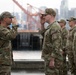 A group of military personnel in OCP (Operational Camouflage Pattern) uniforms are engaged in a formal saluting exchange at what appears to be a waterfront or port location. Two soldiers in the foreground are exchanging salutes, while a line of additional soldiers stands at attention in the background. The setting appears to be an urban port area with cranes and city buildings visible in the background.