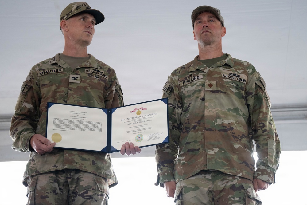 Two U.S. Coast Guard personnel in OCP (Operational Camouflage Pattern) uniforms stand at attention during a military awards ceremony. The individual on the left, identified by name tape as 'KRAUTLER,' holds an open certificate folder displaying a Meritorious Service Medal citation from the United States of America. The individual on the right, identified as 'PHILLIPS,' stands at parade rest beside him.