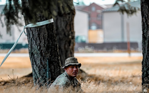 A soldier in camouflage uniform and boonie hat is positioned in a concealed stance behind a tree trunk in a field of dry golden grass. The individual is holding what appears to be an M16/M4-style rifle and appears to be conducting a field training exercise near military-style buildings visible in the background. The scene suggests a tactical training scenario on or near a military installation.